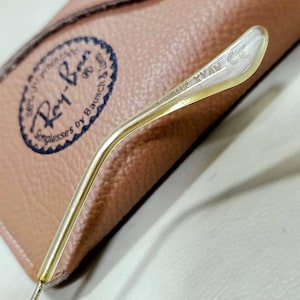May include: A gold-toned metal temple arm from a pair of sunglasses with the text L2802 XVAW CE engraved on the inner side. The arm is positioned over a tan textured case featuring a circular logo with the text Ray-Ban Sunglasses by Bausch & Lomb.