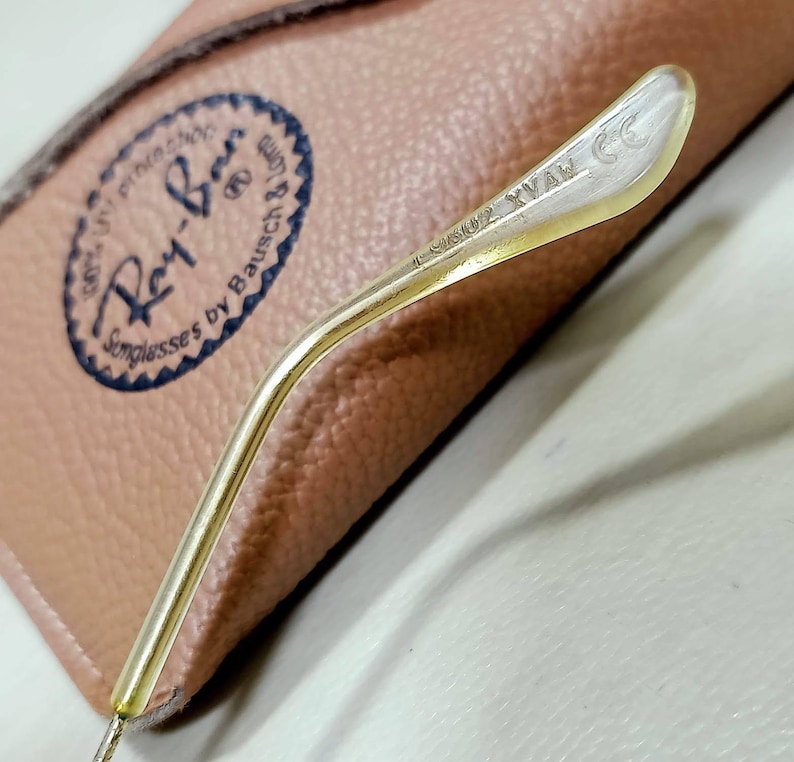 May include: A gold-toned metal temple arm from a pair of sunglasses with the text L2802 XVAW CE engraved on the inner side. The arm is positioned over a tan textured case featuring a circular logo with the text Ray-Ban Sunglasses by Bausch & Lomb.