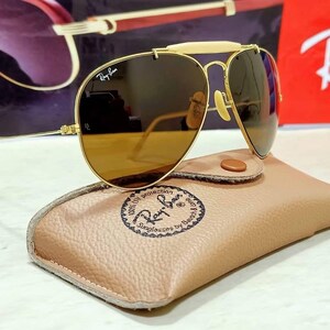 May include: Gold-tone aviator sunglasses featuring a cream-colored brow bar, brown lenses with the Ray-Ban logo, and thin metal temples. The sunglasses rest on a tan textured leather case stamped with the Ray-Ban Sunglasses by Bausch & Lomb logo.