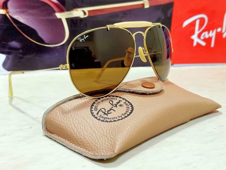 May include: Gold-tone aviator sunglasses featuring a cream-colored brow bar, brown lenses with the Ray-Ban logo, and thin metal temples. The sunglasses rest on a tan textured leather case stamped with the Ray-Ban Sunglasses by Bausch & Lomb logo.