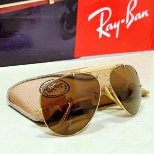May include: Gold-tone aviator sunglasses with brown lenses and a cream-colored brow bar. The sunglasses feature a metal frame and are positioned in front of a tan textured case and a red box with the white Ray-Ban logo printed on the front.