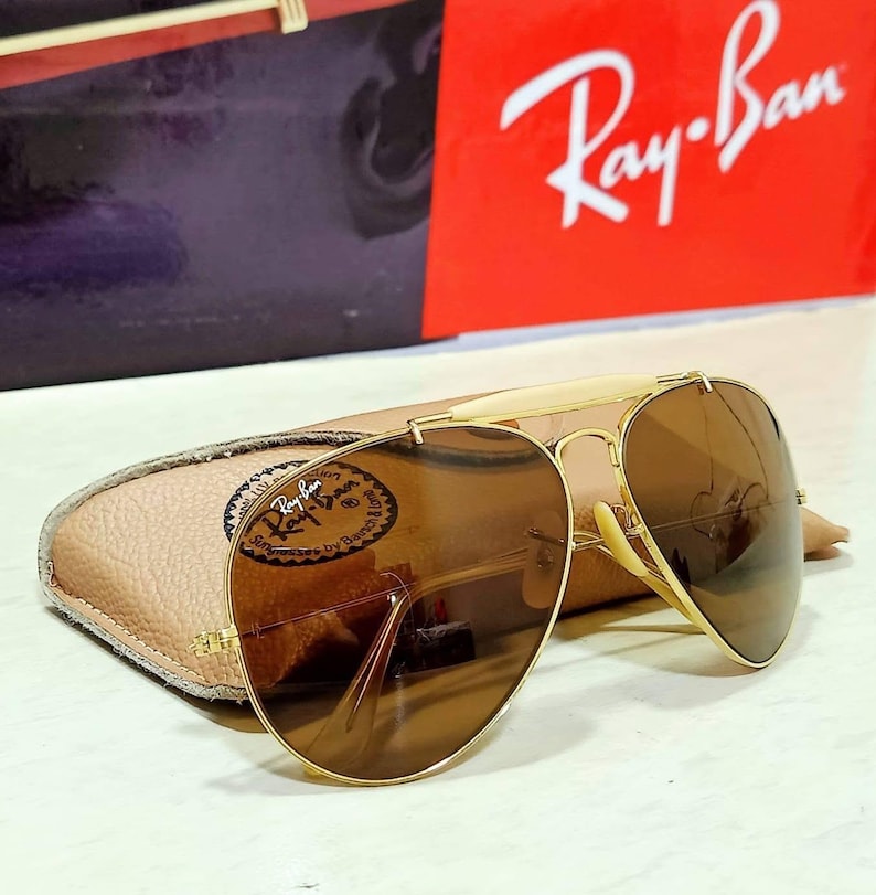 May include: Gold-tone aviator sunglasses with brown lenses and a cream-colored brow bar. The sunglasses feature a metal frame and are positioned in front of a tan textured case and a red box with the white Ray-Ban logo printed on the front.