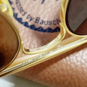 May include: Gold-tone metal sunglasses featuring brown lenses. The bridge is engraved with B&L RAY-BAN 62 14. The background shows a partial logo reading Ray-Ban Sunglasses by Bausch & Lomb. The frame has a metallic finish and a double bridge design.