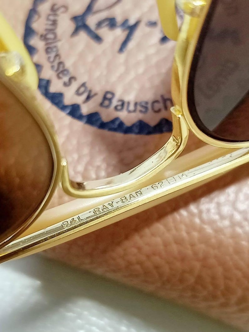 May include: Gold-tone metal sunglasses featuring brown lenses. The bridge is engraved with B&L RAY-BAN 62 14. The background shows a partial logo reading Ray-Ban Sunglasses by Bausch & Lomb. The frame has a metallic finish and a double bridge design.