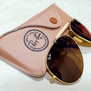 May include: Gold-tone aviator sunglasses with brown lenses and the Ray-Ban logo on the lens. The sunglasses are paired with a tan, textured leather-like carrying case featuring a circular Ray-Ban logo stamp and a single brown snap button closure.