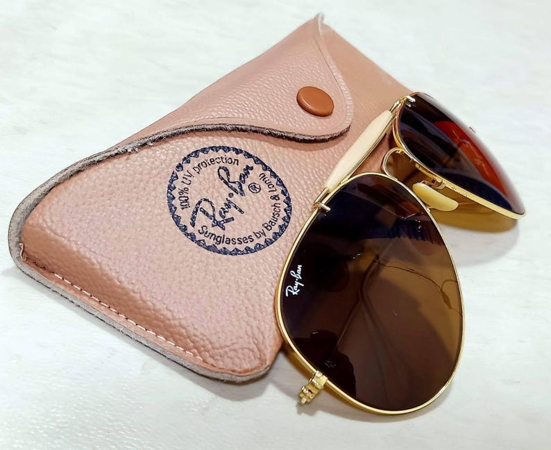 May include: Gold-tone aviator sunglasses with brown lenses and the Ray-Ban logo on the lens. The sunglasses are paired with a tan, textured leather-like carrying case featuring a circular Ray-Ban logo stamp and a single brown snap button closure.