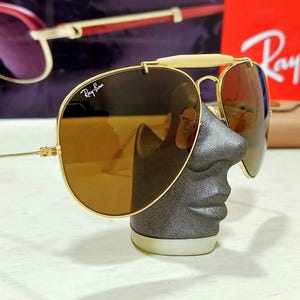 May include: Gold-tone aviator sunglasses featuring brown lenses and a white brow bar. The Ray-Ban logo is printed in white on the upper right lens. The thin metal frame includes adjustable nose pads and slender temples, showcasing a classic design.
