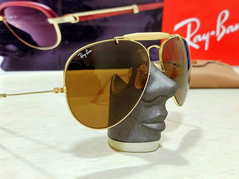 May include: Gold-tone aviator sunglasses featuring brown lenses and a white brow bar. The Ray-Ban logo is printed in white on the upper right lens. The thin metal frame includes adjustable nose pads and slender temples, showcasing a classic design.