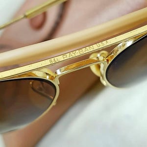 May include: Gold-tone metal sunglasses featuring a brow bar with the engraved text B&L RAY-BAN U.S.A. The frames have thin gold-tone metal temples with light beige plastic temple tips and dark brown tinted lenses, showcasing a classic aviator design.