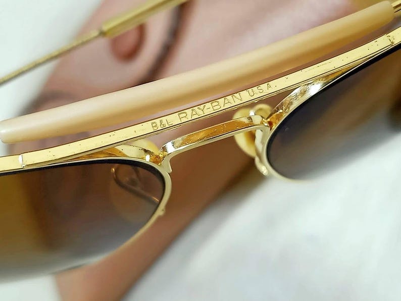 May include: Gold-tone metal sunglasses featuring a brow bar with the engraved text B&L RAY-BAN U.S.A. The frames have thin gold-tone metal temples with light beige plastic temple tips and dark brown tinted lenses, showcasing a classic aviator design.