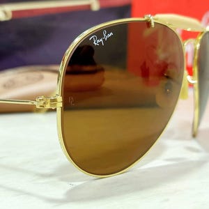 May include: Gold-tone aviator sunglasses featuring brown-tinted lenses with the white Ray-Ban logo printed on the upper right lens and the letters BL etched near the hinge. The thin metal frame includes a double bridge and adjustable nose pads.