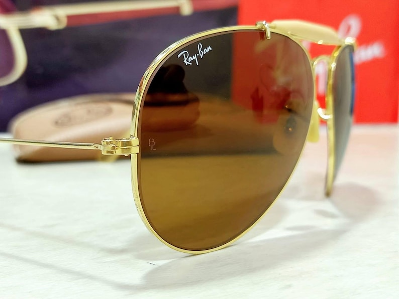 May include: Gold-tone aviator sunglasses featuring brown-tinted lenses with the white Ray-Ban logo printed on the upper right lens and the letters BL etched near the hinge. The thin metal frame includes a double bridge and adjustable nose pads.