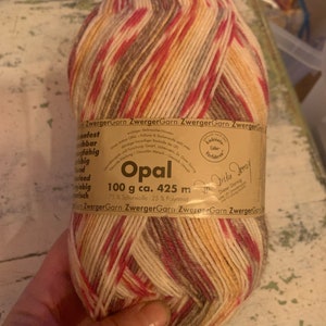 May include: A ball of yarn in shades of beige, brown, pink, and red. The yarn is labeled "Opal" and is 100 grams and 425 meters long. It is made of 75% wool and 25% polyamide.