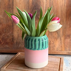 May include: A colourful ceramic vase in light green, pink, and cream, filled with tulips. The tulips are pink, white, and purple. The vase sits on a wooden chopping board. The vase has a height of approximately 15 cm.