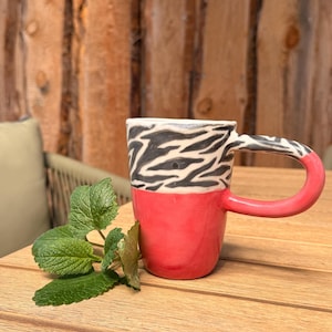 Könnte beinhalten: Handgefertigte Keramiktasse mit leuchtend rotem Boden und Griff. Der obere Teil zeigt ein schwarz-weißes Zebramuster. Die Tasse steht auf einem Holztisch, daneben grüne Blätter.