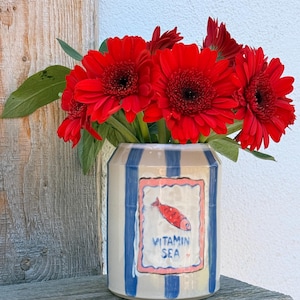 Peut inclure: Un vase en céramique à rayures bleues et blanches contient un bouquet de gerberas rouges vifs. Le vase présente une étiquette rectangulaire avec une illustration de poisson rouge et les mots "VITAMIN SEA". Le vase est sur une surface en bois.