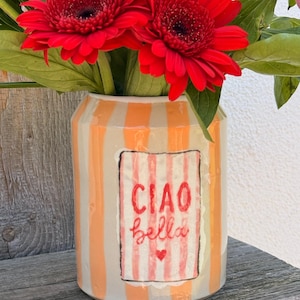 Könnte beinhalten: Eine Keramikvase mit orangefarbenen und weißen Streifen, gefüllt mit leuchtend roten Gerbera-Gänseblümchen. Die Vase hat ein rechteckiges Etikett mit dem Schriftzug "CIAO bella" in roter Schrift und einem kleinen Herz.