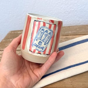 Op de afbeelding: Een keramieken beker met een handgeschilderd ontwerp van een sardineblikje op een achtergrond van verticale rode en witte strepen. De illustratie bevat drie blauwe vissen en het woord SARDINE in blauwe inkt op de voorzijde van de beker.