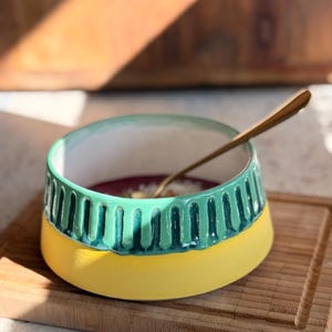 Peut inclure: Un bol en céramique avec une base jaune et une section supérieure turquoise avec des rainures verticales. Le bol contient un mélange violet et une cuillère dorée. Le bol est posé sur une planche à découper en bois.