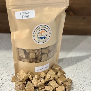 May include: A brown paper bag of freeze-dried lamb liver for pets. The bag has a clear window showing the contents and a label that reads "Lamb Liver." Cubed pieces of lamb liver are scattered around the bag.