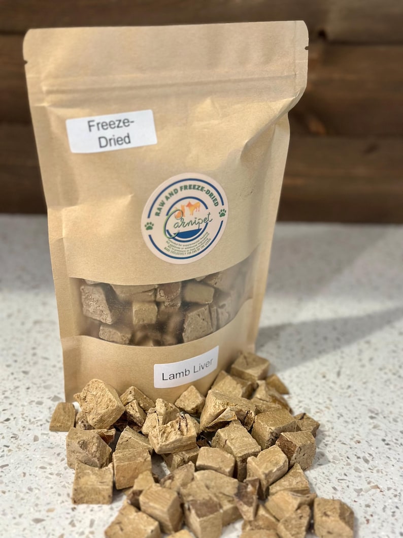 May include: A brown paper bag of freeze-dried lamb liver for pets. The bag has a clear window showing the contents and a label that reads "Lamb Liver." Cubed pieces of lamb liver are scattered around the bag.