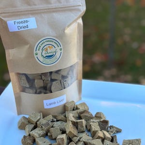 May include: A brown paper bag of freeze-dried lamb liver for pets, with a clear window showing the cubed treats. The bag has a label that says "Lamb Liver" and a logo that says "Raw and Freeze-Dried". Cubed treats are scattered on a white surface.