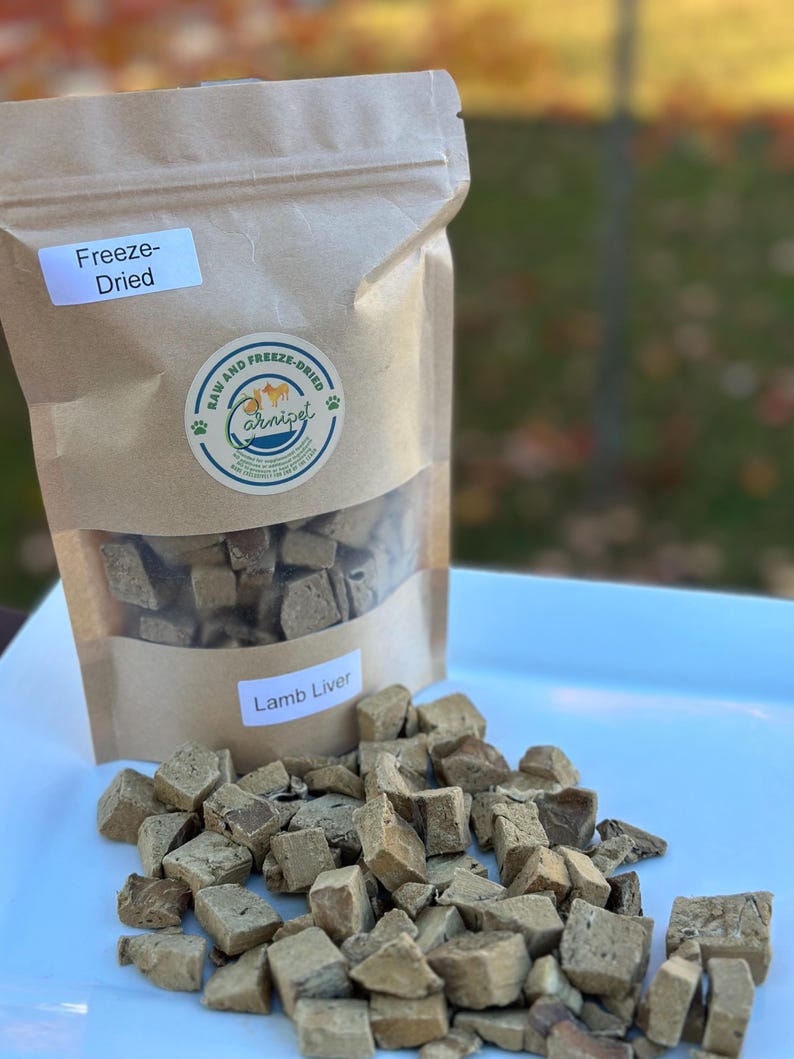 May include: A brown paper bag of freeze-dried lamb liver for pets, with a clear window showing the cubed treats. The bag has a label that says "Lamb Liver" and a logo that says "Raw and Freeze-Dried". Cubed treats are scattered on a white surface.