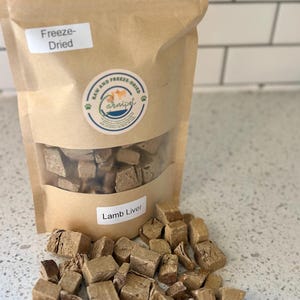 May include: A brown paper bag of freeze-dried lamb liver for pets. The bag has a clear window and a label that reads "Lamb Liver." Cubed pieces of the product are scattered in front of the bag. The bag also has a round sticker with the words "Raw and Freeze-Dried" and a paw print.