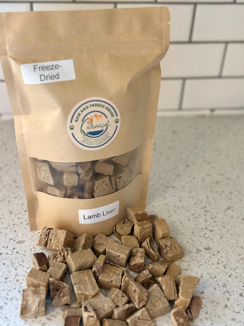 May include: A brown paper bag of freeze-dried lamb liver for pets. The bag has a clear window and a label that reads "Lamb Liver." Cubed pieces of the product are scattered in front of the bag. The bag also has a round sticker with the words "Raw and Freeze-Dried" and a paw print.