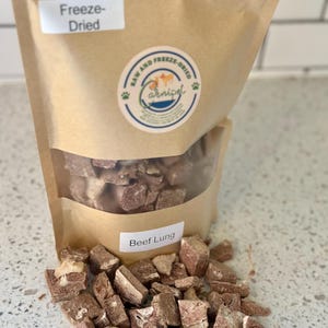 May include: A brown paper bag of freeze-dried beef lung for pets. The bag has a clear window showing the contents and a label that reads "Beef Lung." A logo with the words "Raw and Freeze-Dried" and the brand name "Carnipel" is also visible.