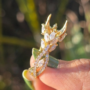 May include: A gold ring with a distinctive design, showcasing marquise-cut and round-cut clear gemstones. The ring has a floral or leaf-like pattern with a curved band. It is held against a blurred green backdrop.