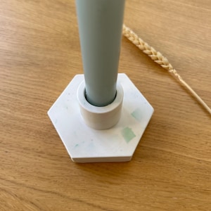 Peut inclure: Une bougie vert clair dans un bougeoir hexagonal blanc avec des éclats verts. Le bougeoir a une petite base circulaire pour la bougie. Un seul brin de blé est en arrière-plan.