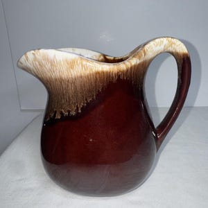 Puede incluir: Una jarra de cerámica esmaltada marrón y blanca con una boca ancha y un cuerpo redondeado. La jarra tiene un esmalte marrón con vetas blancas y un borde blanco.