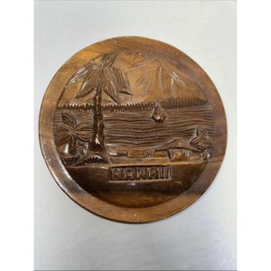 Op de afbeelding: Een ronde houten snijwerk met een Hawaïaanse strandscène. Het snijwerk toont palmbomen, een zeilboot en het woord "HAWAII" onderaan. Het hout heeft een warme, bruine tint.