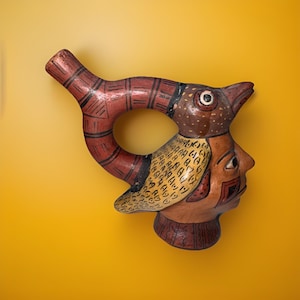 May include: A handcrafted ceramic vessel featuring a bird-like head and a human profile. The vessel is painted with earthy tones of red, brown, and yellow, with intricate patterns and designs. The unique shape and detailed artistry make it a decorative piece.
