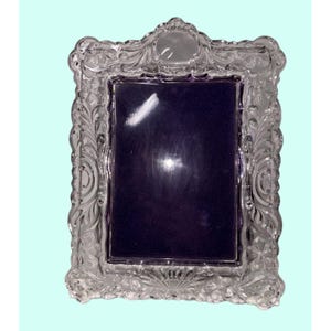 Puede incluir: Un marco de fotos transparente y ornamentado con un respaldo morado oscuro. El marco presenta intrincados diseños florales y de volutas, con una parte superior redondeada y una abertura rectangular para una fotografía. El marco está hecho de un material transparente, posiblemente vidrio o cristal.