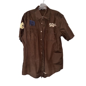 May include: Brown short-sleeved button-up shirt with two front pockets. Features patches on the left sleeve and chest, and embroidered text that reads "Des Gants G. Essen Festival 1972". The shirt is hanging on a clear hanger.