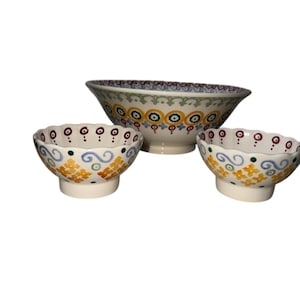 May include: A set of three ceramic bowls with a cream-colored base and hand-painted patterns. The large bowl features a repeating yellow and blue circular design, while the two smaller bowls have scalloped edges with yellow floral motifs.