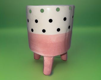 Może przedstawiać: Mała ceramiczna doniczka. Górna część jest biała z czarnymi kropkami, dolna część jest różowa. Ma trzy różowe nóżki i otwory drenażowe w pobliżu góry. Przedmiot dekoracyjny.