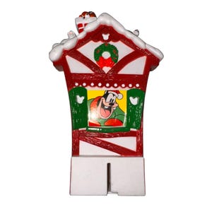 Op de afbeelding: Een rood en wit kerstversieringshuis met Goofy met een kerstmuts. Het huis heeft groene luiken, een krans en een schoorsteen met een klein cadeau. Sneeuw accentueert het dak.
