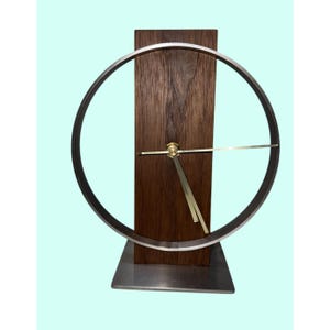May include: A table clock featuring a vertical dark brown wood plank mounted on a square metal base. A large silver circular frame surrounds the wood, with thin gold-coloured clock hands attached to the centre of the wood plank.
