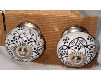 Könnte beinhalten: Zwei weiße Keramik-Schrankknöpfe mit schwarzen Blumenmustern. Jeder Knopf hat einen silberfarbenen Sockel und eine dekorative silberne Mitte. Die Knöpfe werden auf einer braunen Kartonrückwand präsentiert.