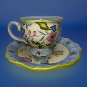 May include: A ceramic teacup and saucer set. The cup and saucer are cream-colored with a floral design in pink, blue, and green. The rim of the cup and saucer is light blue with a green trim. The set is on a blue background.