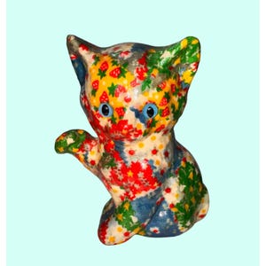 May include: A colorful ceramic cat figurine with blue eyes and a raised paw. The cat is decorated with a pattern of red strawberries, yellow flowers, and green leaves on a white and blue background.