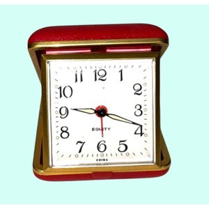 May include: A square travel alarm clock with a red folding case and a gold-toned metal frame. The white clock face features black Arabic numerals, black hour and minute hands with luminous centers, a thin red second hand, and the text Equity and China.