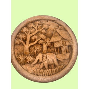 May include: A round, wooden carving depicting an elephant walking through a landscape with trees and a building. The relief carving is in shades of brown and tan, with detailed textures and patterns.