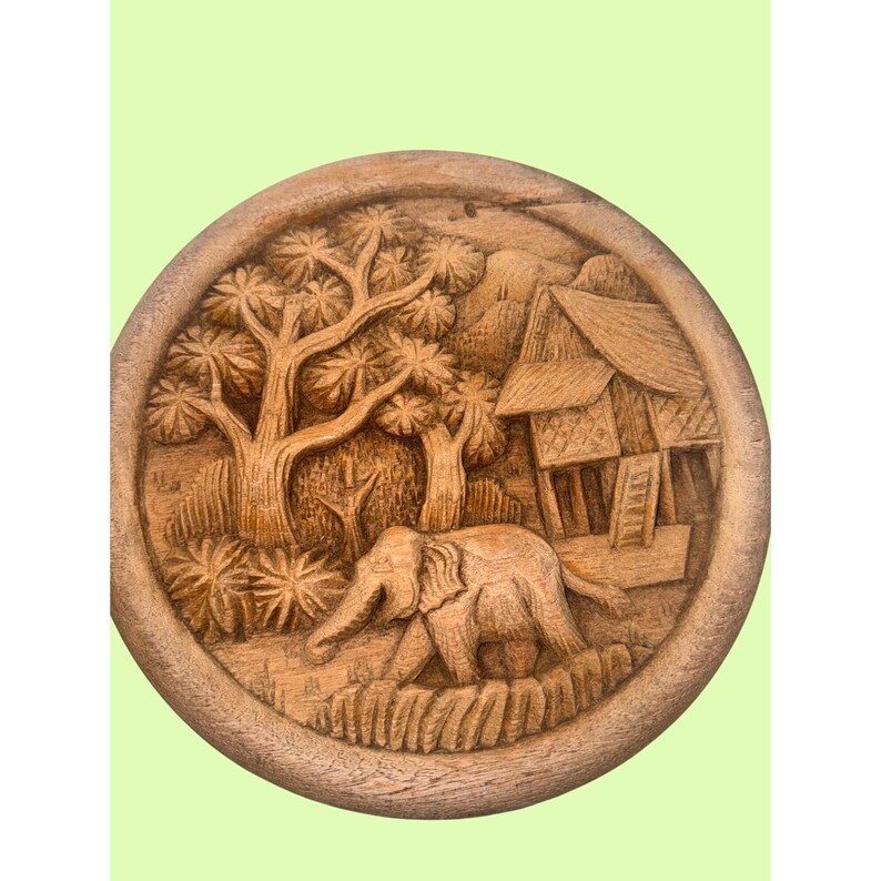 May include: A round, wooden carving depicting an elephant walking through a landscape with trees and a building. The relief carving is in shades of brown and tan, with detailed textures and patterns.