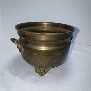 Pode incluir: Um vaso de latão com duas alças e três pés. O vaso tem um aspeto desgastado e envelhecido.