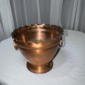 May include: A copper-coloured metal bowl with a wide, scalloped rim and a circular pedestal base. The bowl features two small, rounded metal handles attached to opposite sides of the exterior, providing a functional grip for this decorative vessel.