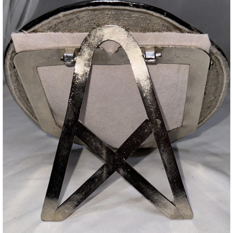 May include: Silver-toned oval picture frame with a light gray backing. The frame has a decorative edge and a metal easel back for display. The frame is designed to hold a small photograph or artwork.